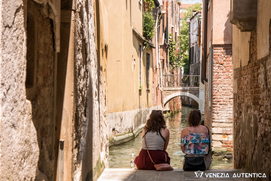 Two tourists in Venice sitting on the edge of a calle, looking at a canal and a bridge in front of them, on a warm summer day.