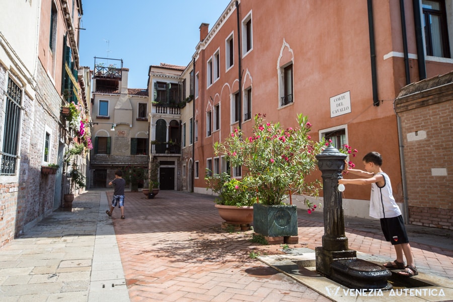 Two young children playing in corte del cavallo. In the foreground a child filling a water balloon at a fountain close to an oleandrum tree with pink flowers. The other child is halfway through the courtyard, close to a balcony filled with flowers.