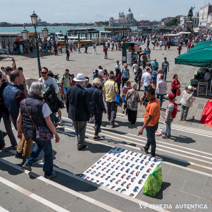 illegal street vendors on a bridge trying to sell glasses and selfie sticks to passers-by. In the background the Salute church, the equestrian statue of Vittorio Emanuele di Savoia and the Doge palace.