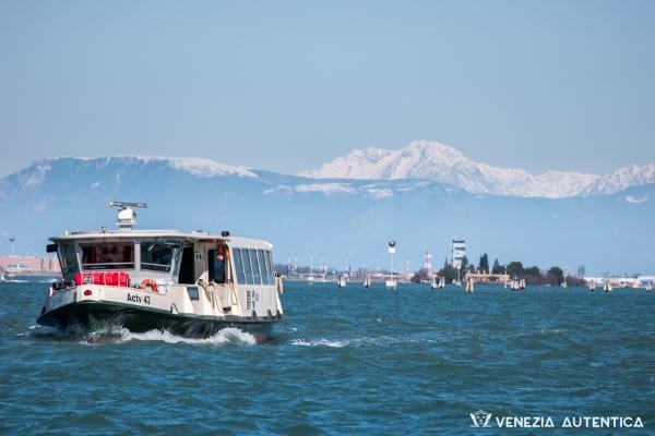 A public water bus navigating in front of the fondamenta nuove on a clear day, with snowy dolomites in the background.